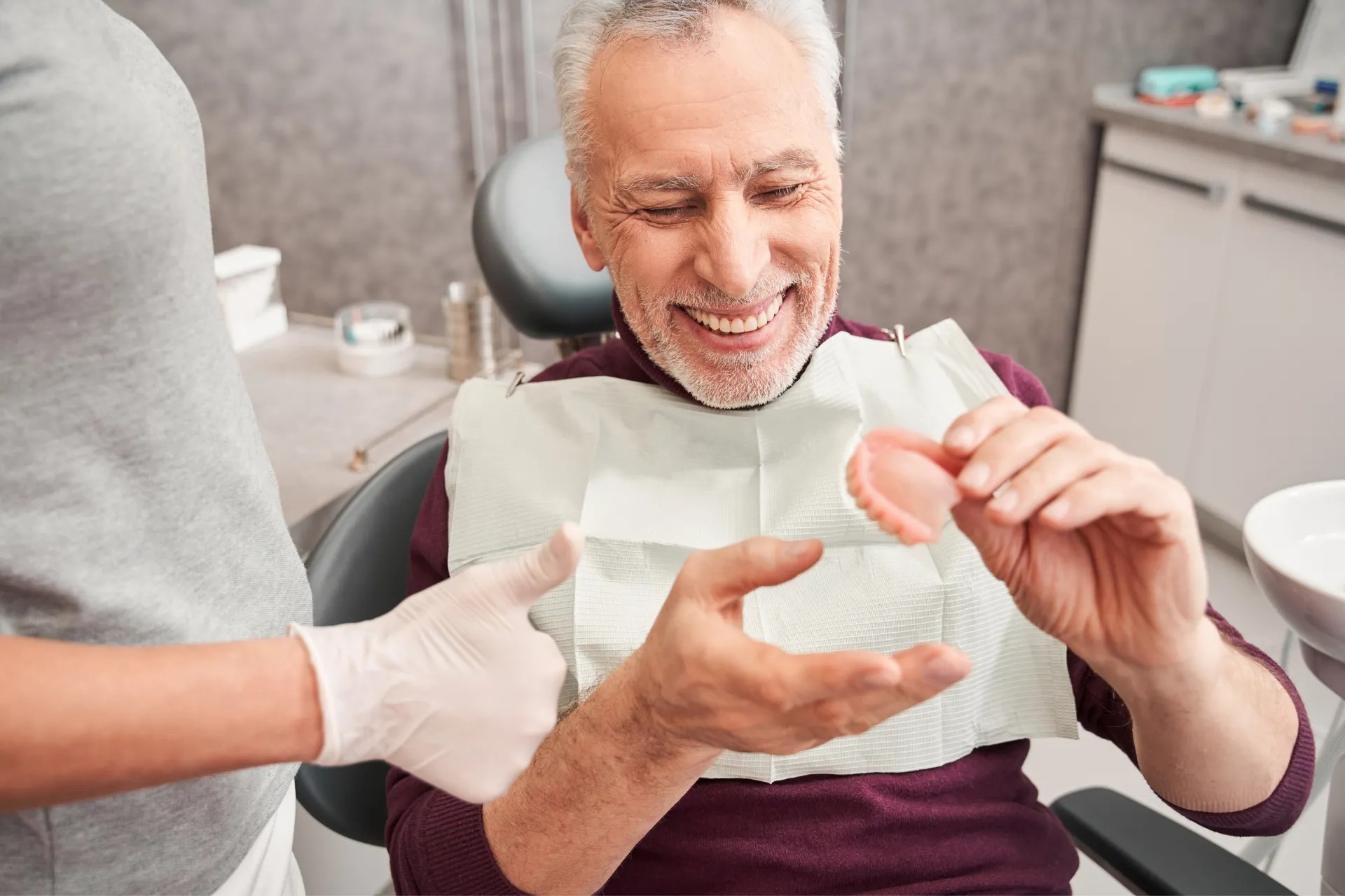 Smiling elderly man sitting in a dental chair holding a set of dentures while a person wearing gloves gives a thumbs-up.