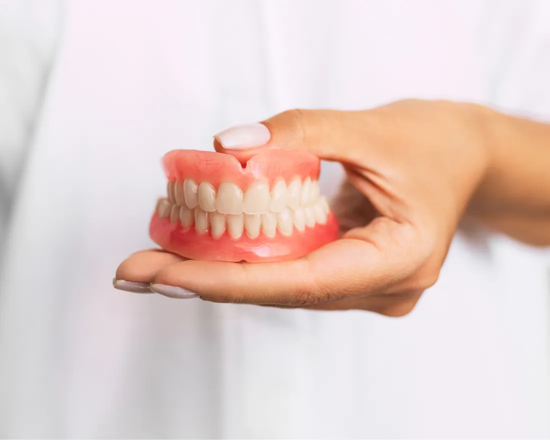 Hand holding a full set of pink and white dentures against a blurred background.