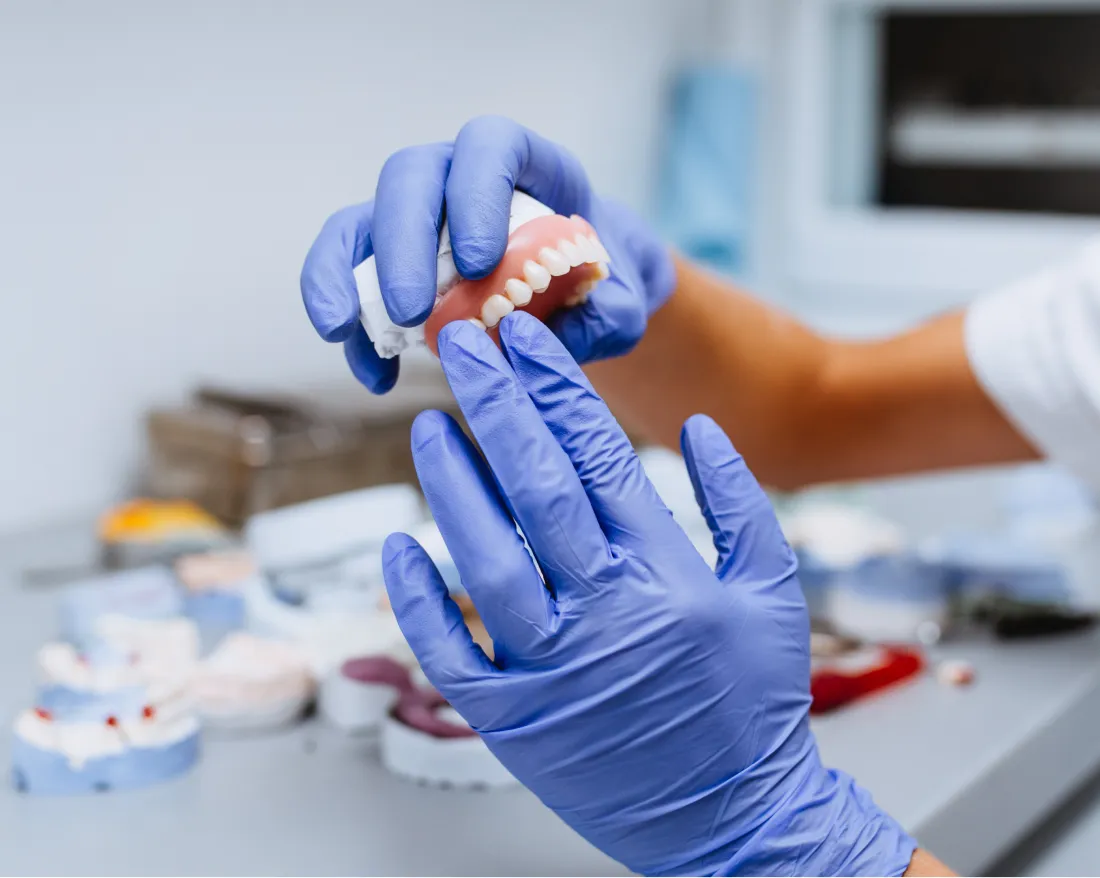 Hands wearing blue gloves holding a dental prosthetic model above a workspace with dental casts.