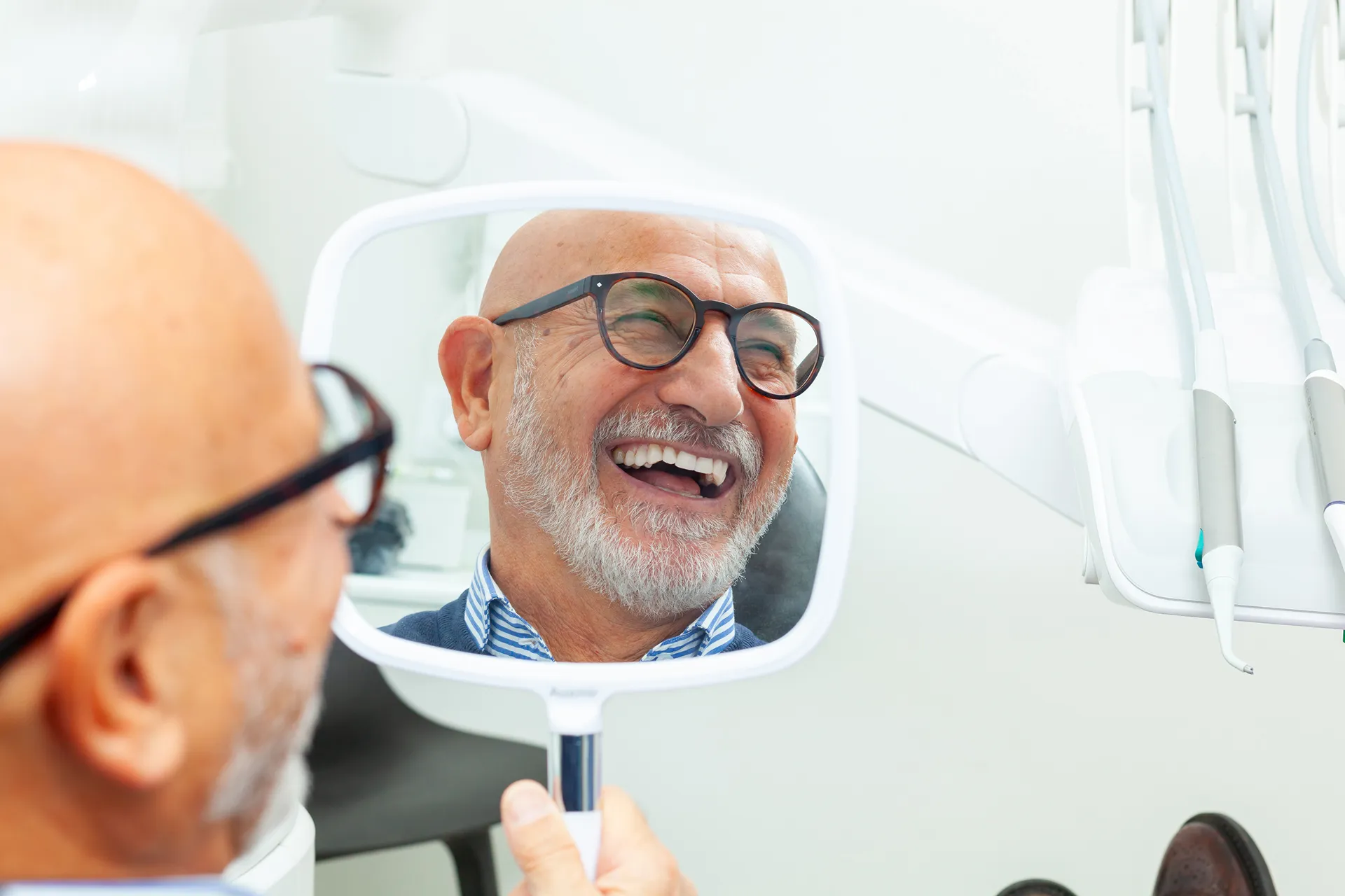 Smiling elderly man with glasses and beard looking at his reflection in a handheld mirror at a dental clinic.