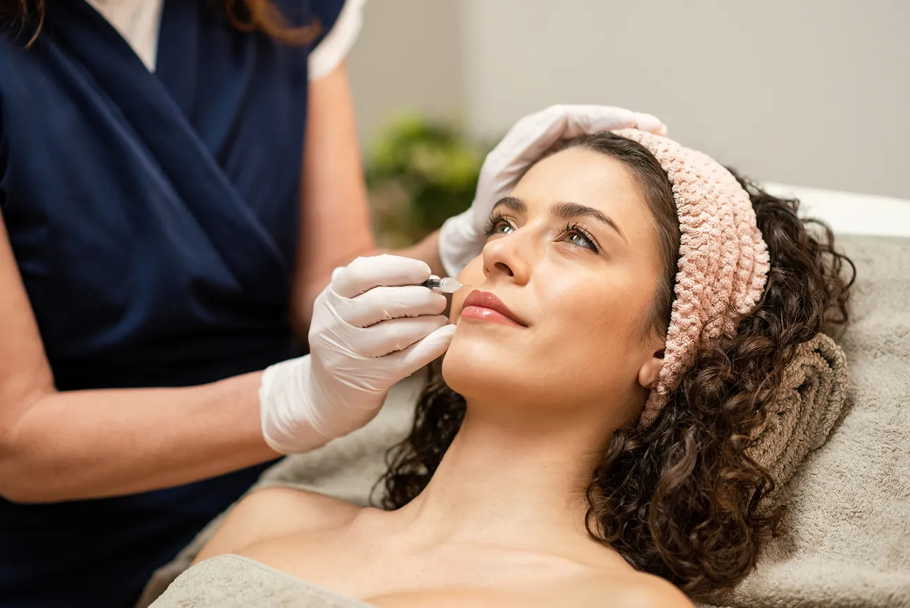 Woman lying down with a headband receiving a lip filler injection from a gloved practitioner.