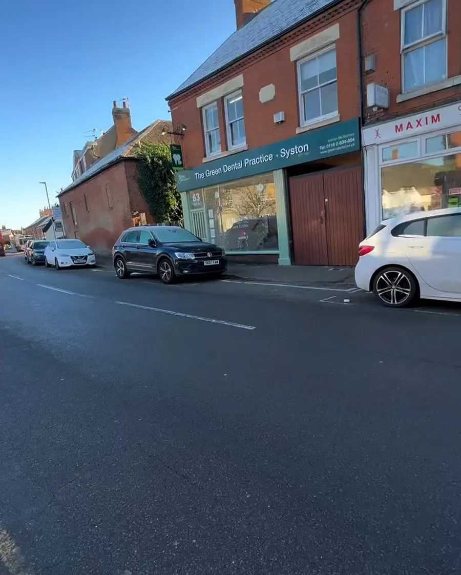 Street view of parked cars along a road with a brick building housing The Green Dental Practice in Syston under a clear blue sky.