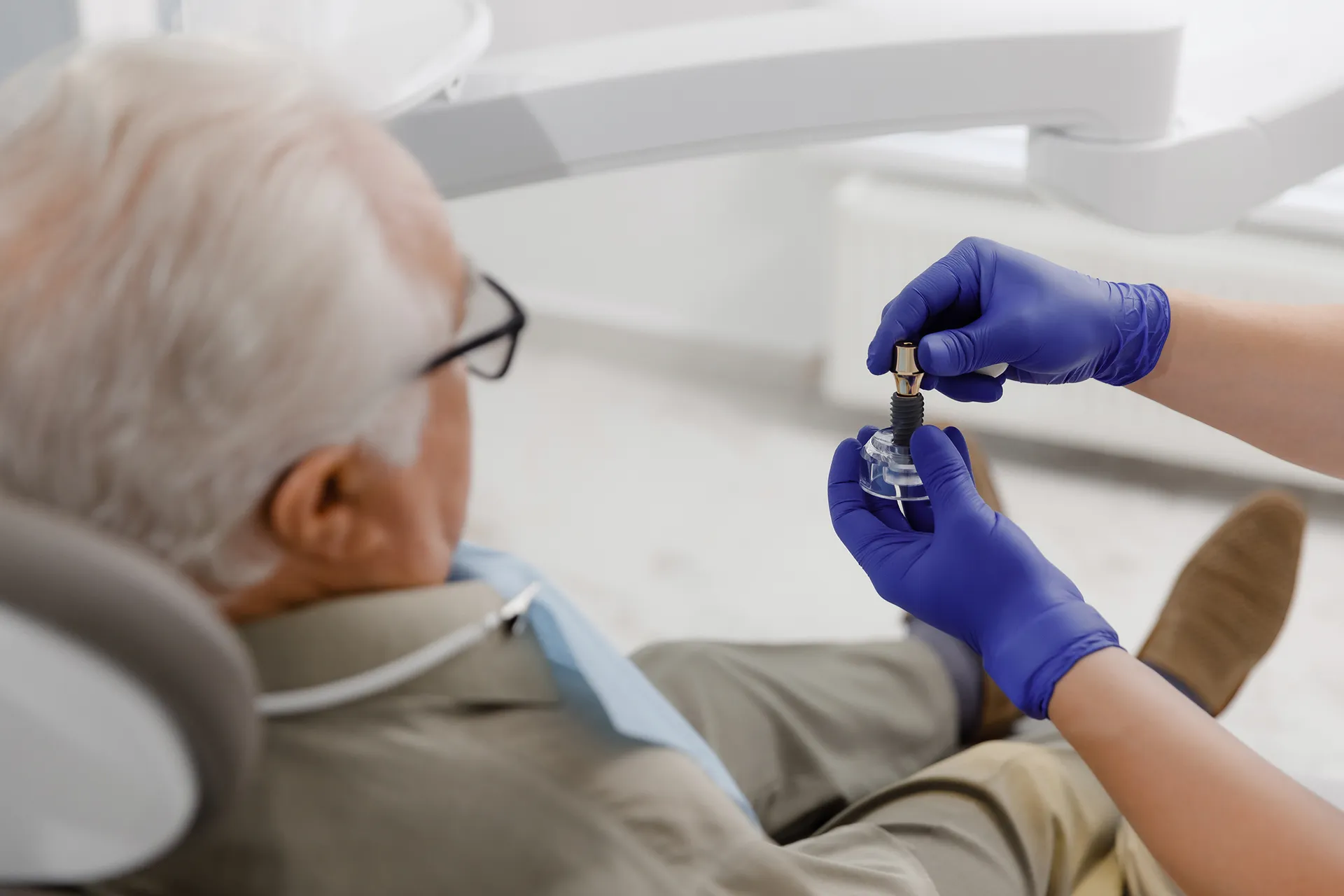 Close-up of a dental professional wearing purple gloves holding a dental implant model near an elderly patient seated in a dental chair.