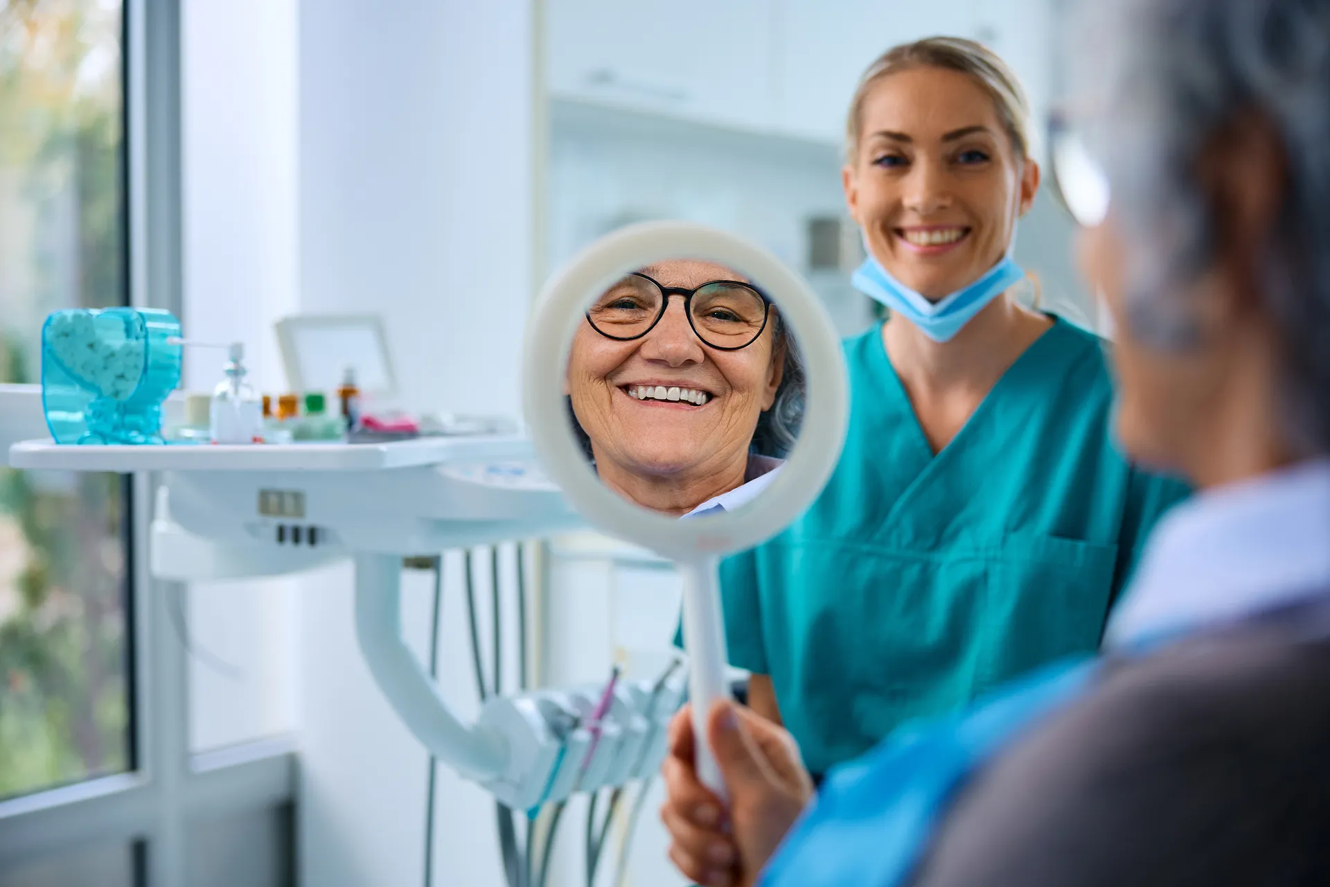 Smiling elderly woman looking at her reflection in a handheld mirror held by a person in a dental office with a female dentist smiling in the background.