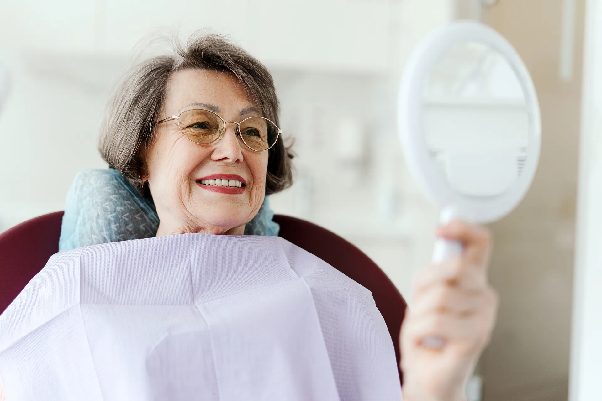 Smiling elderly woman wearing glasses sitting in a dental chair holding a hand mirror.