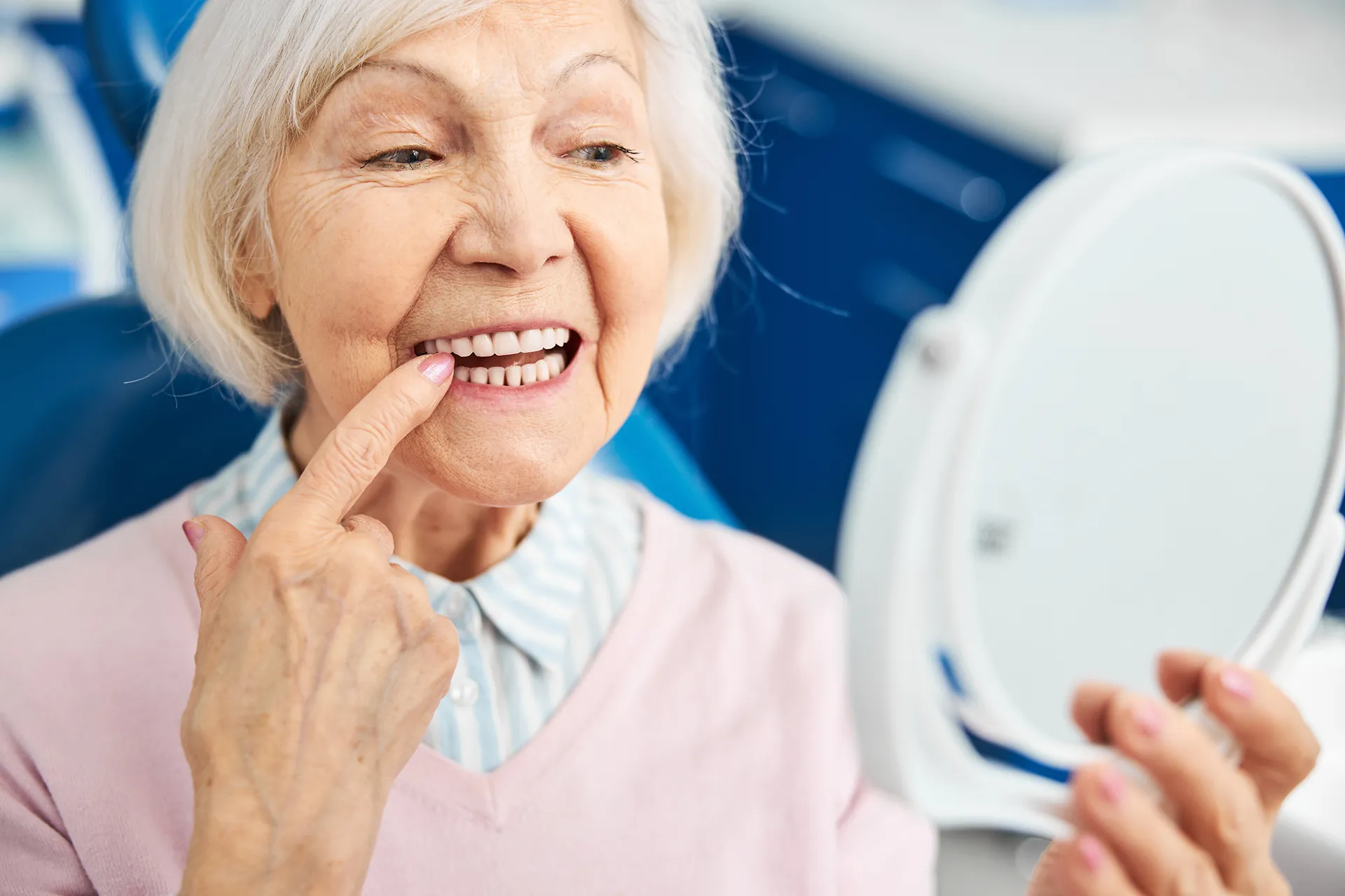 Elderly woman smiling and pointing to her teeth while holding a mirror in a dental clinic.