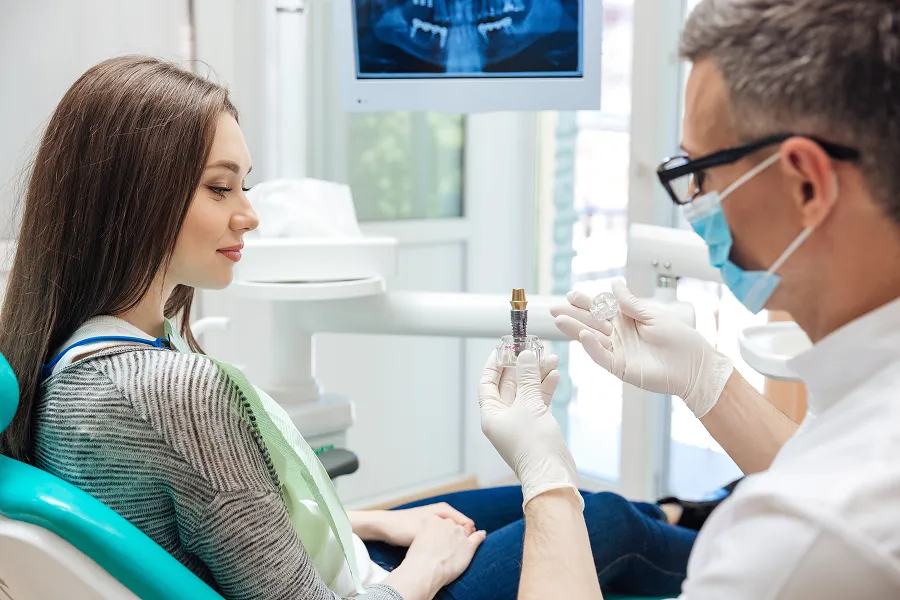 Dentist wearing gloves and mask showing a dental implant to a seated female patient in a dental office.