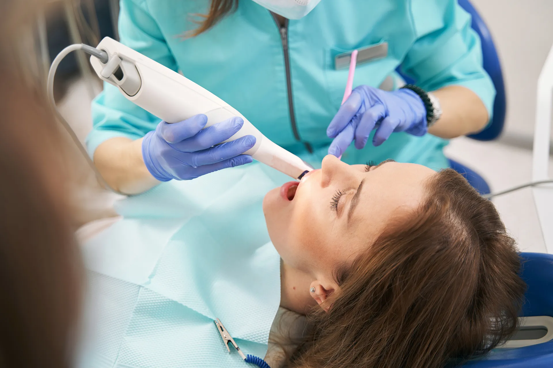 Dentist wearing blue gloves using a dental scanner inside the mouth of a female patient reclining in a dental chair.