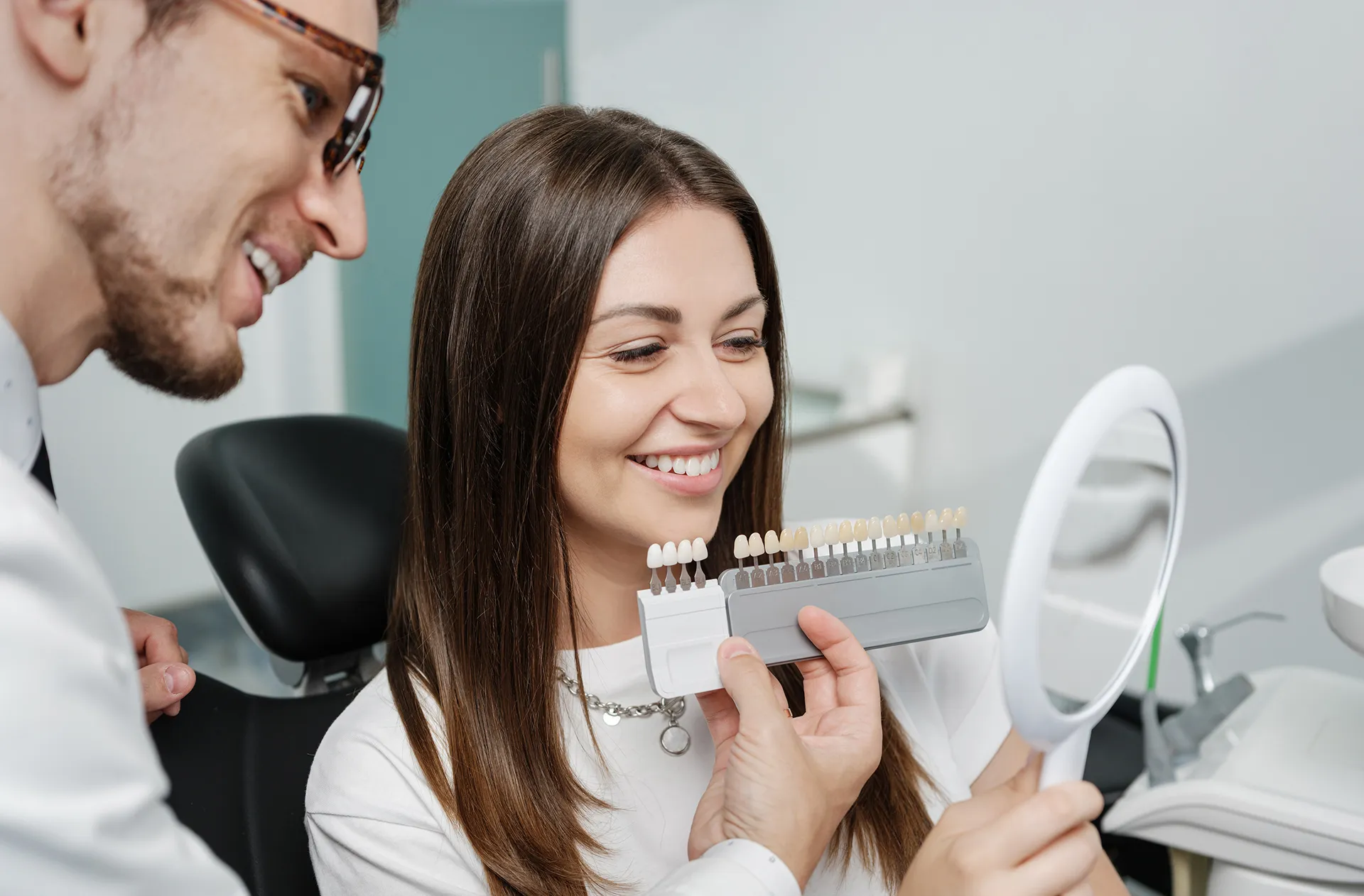 Smiling woman holding a mirror while a dentist compares dental veneer shades to her teeth in a dental office.
