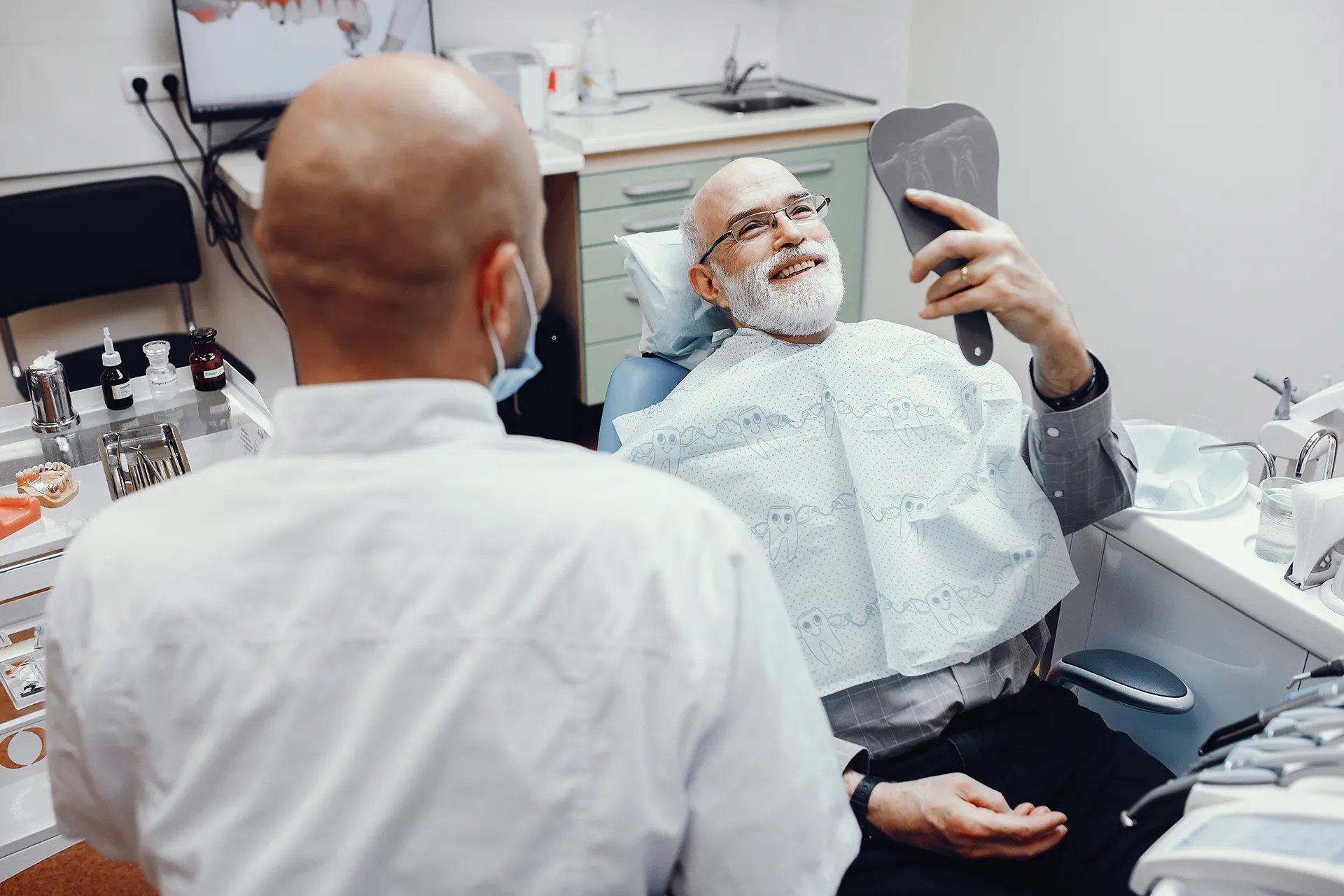 Smiling elderly man sitting in a dental chair holding a mirror while a dentist stands nearby.