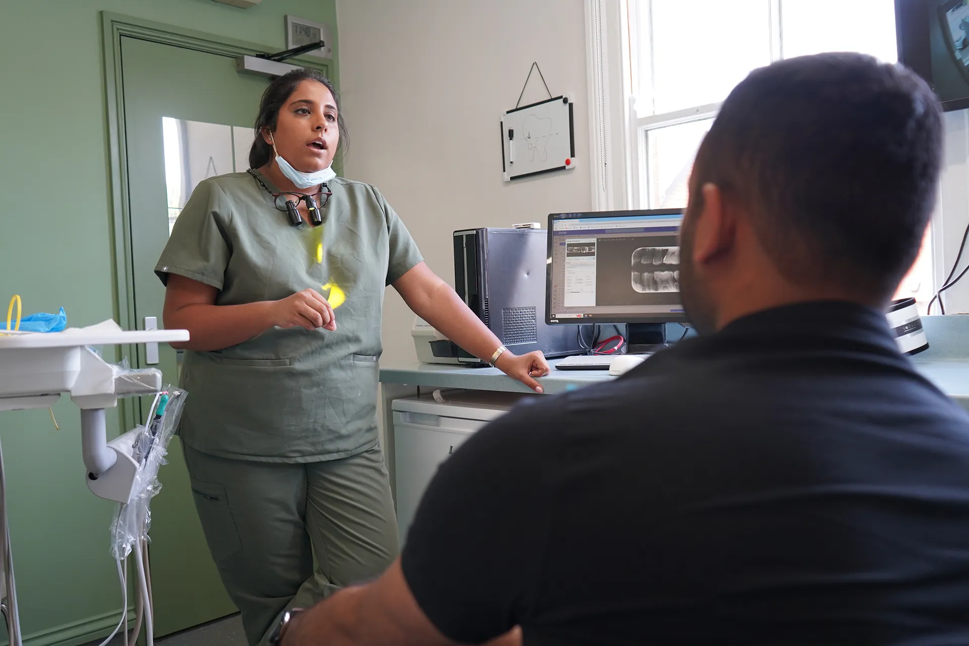 Dentist in green scrubs explaining dental X-ray displayed on a computer screen to a seated patient.