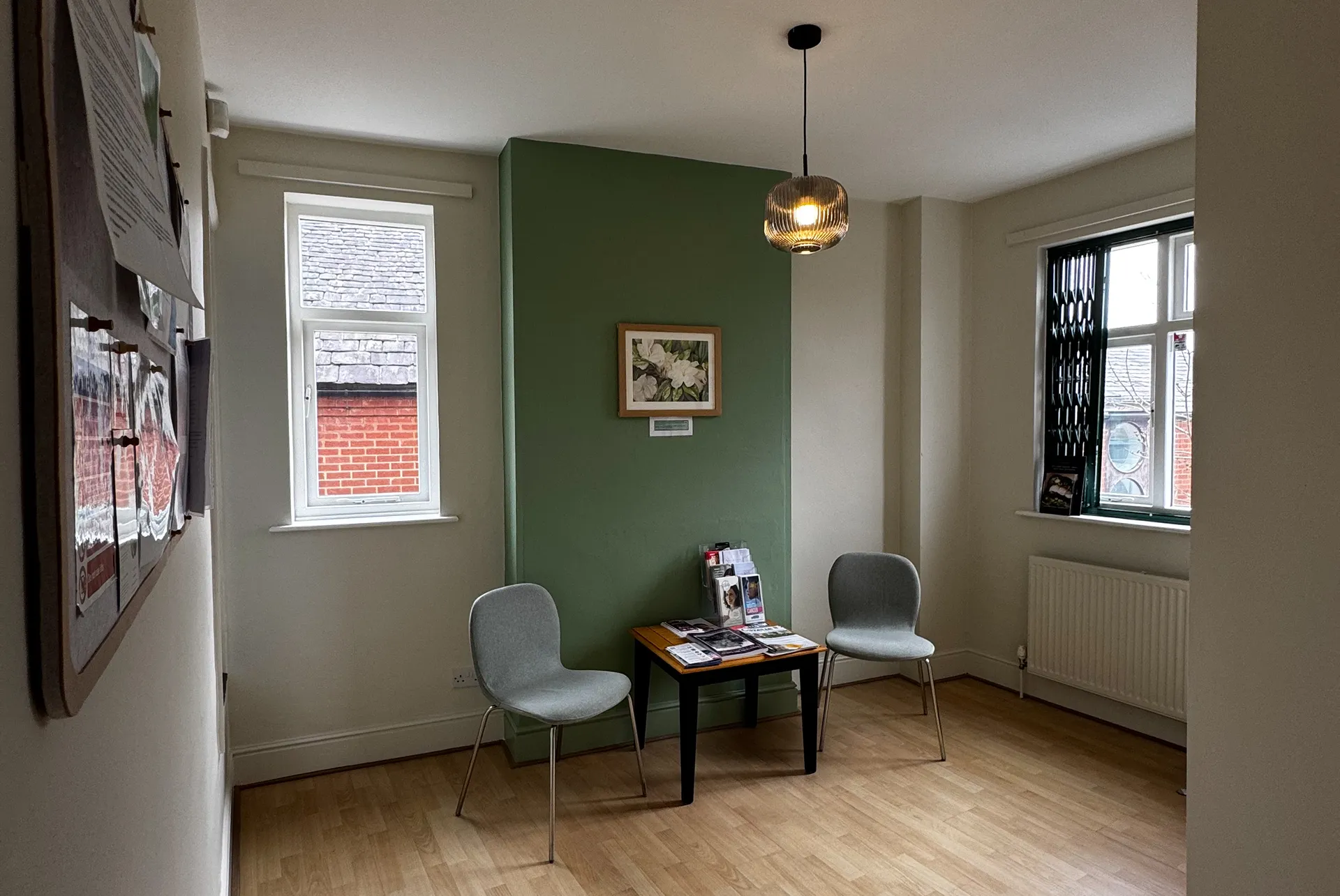 Small waiting area with two gray chairs, a wooden table holding magazines, a green accent wall with a framed floral picture, and two windows.
