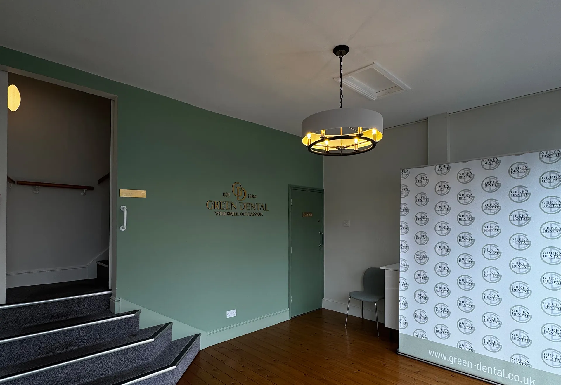 Reception area of Green Dental with green walls, wooden floor, staircase, hanging light fixture, and branded banner.