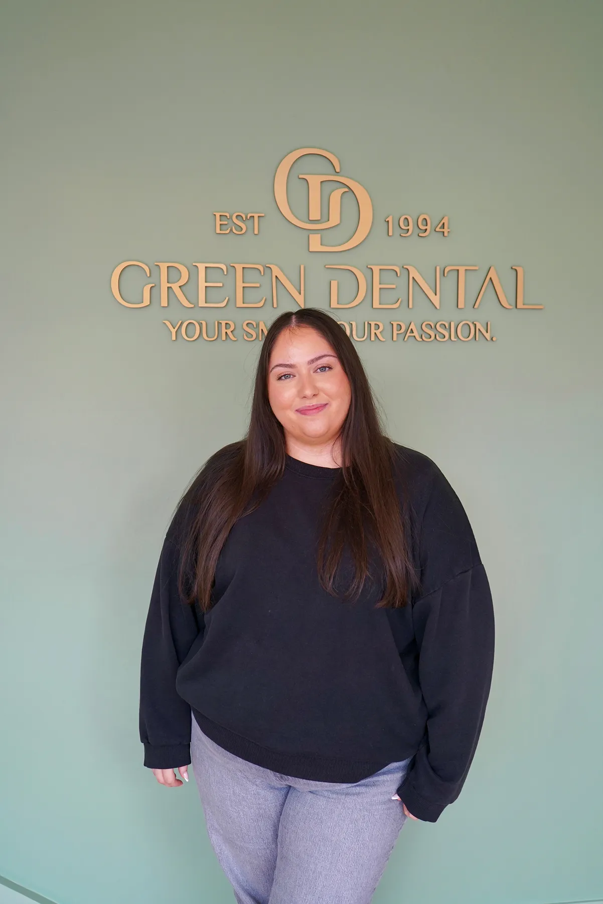 Smiling woman with long brown hair wearing a black sweater and gray pants standing in front of a green wall with Gold Green Dental logo and slogan.