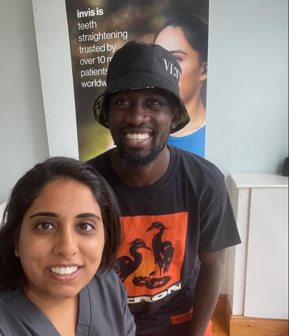 Smiling man wearing a black bucket hat and a woman with dark hair taking a selfie indoors with a poster about teeth straightening in the background.