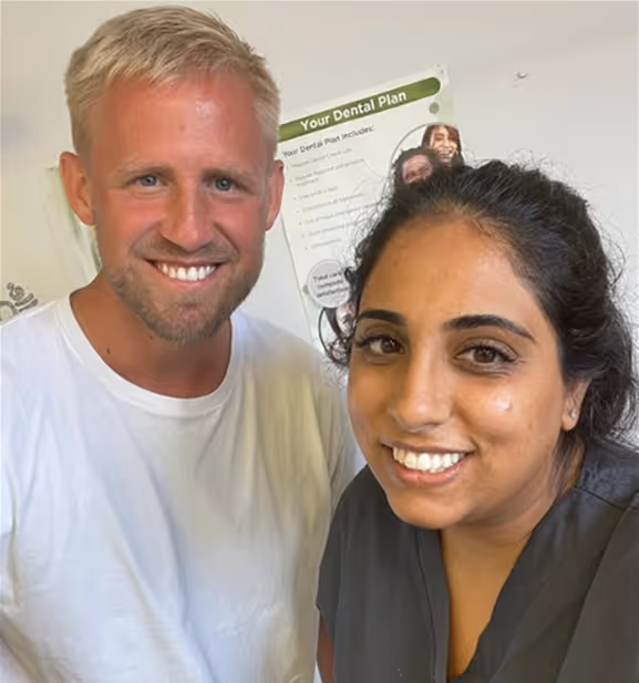 Smiling man in a white shirt and woman in a dark top posing for a close-up photo indoors with a dental plan poster in the background.