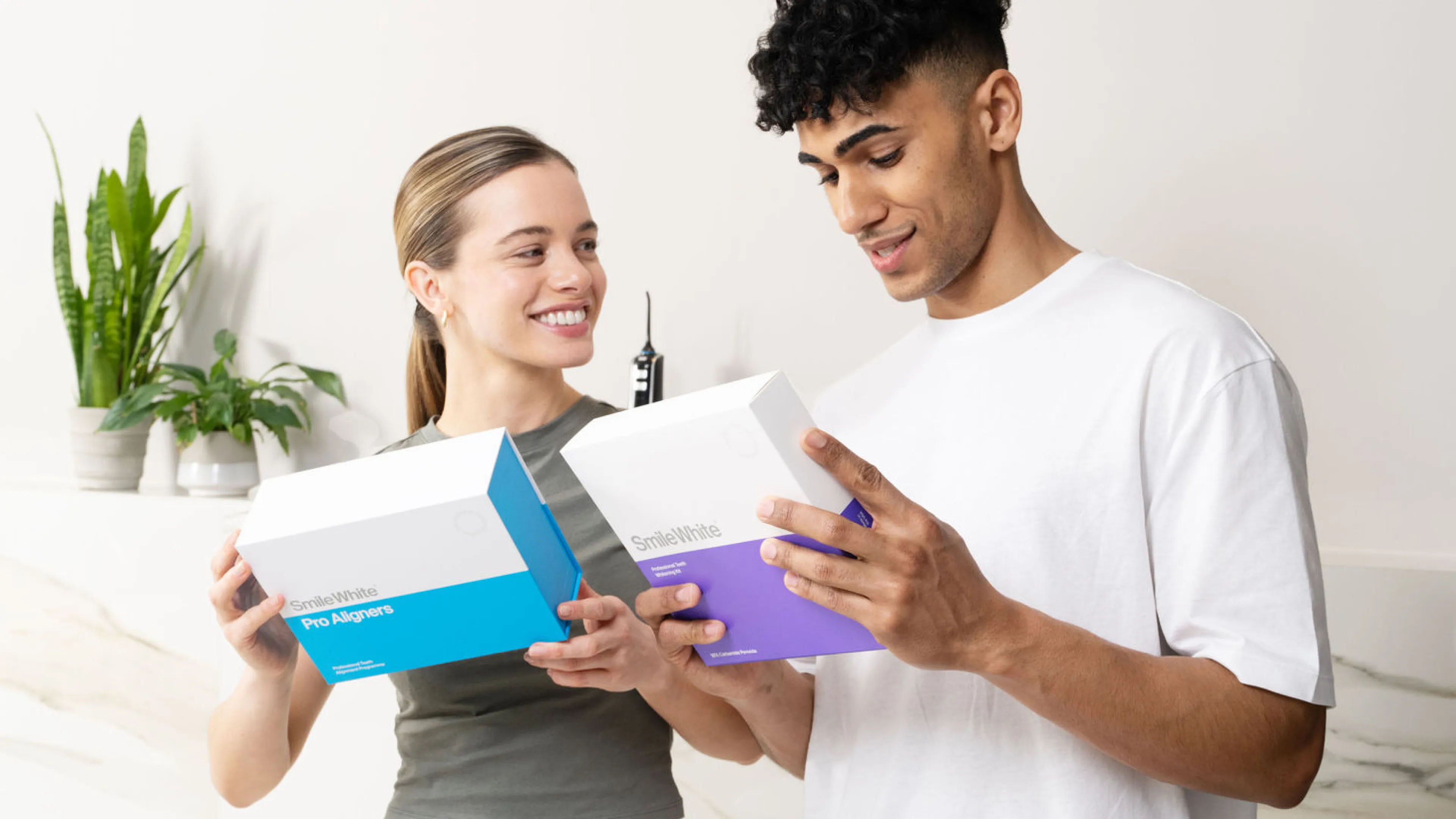 Smiling man and woman each holding a SmileWhite product box, standing indoors with plants in the background.