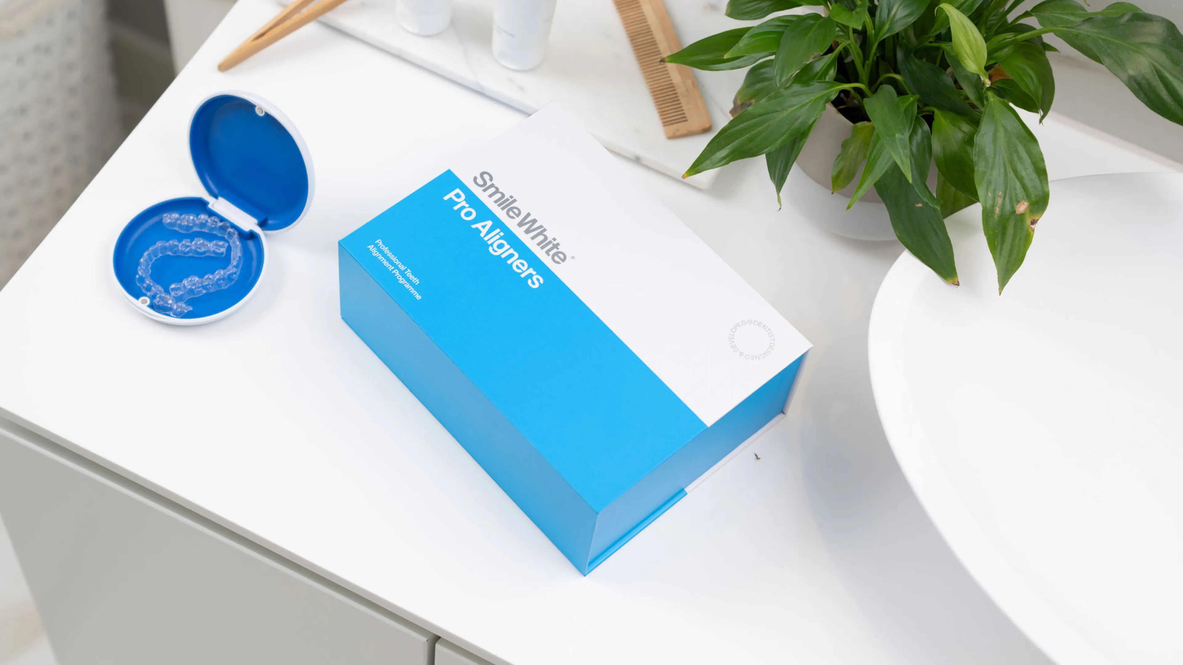 Smile White Pro Aligners box on a white counter next to an open blue case with clear dental aligners, a wooden comb, and a green potted plant.