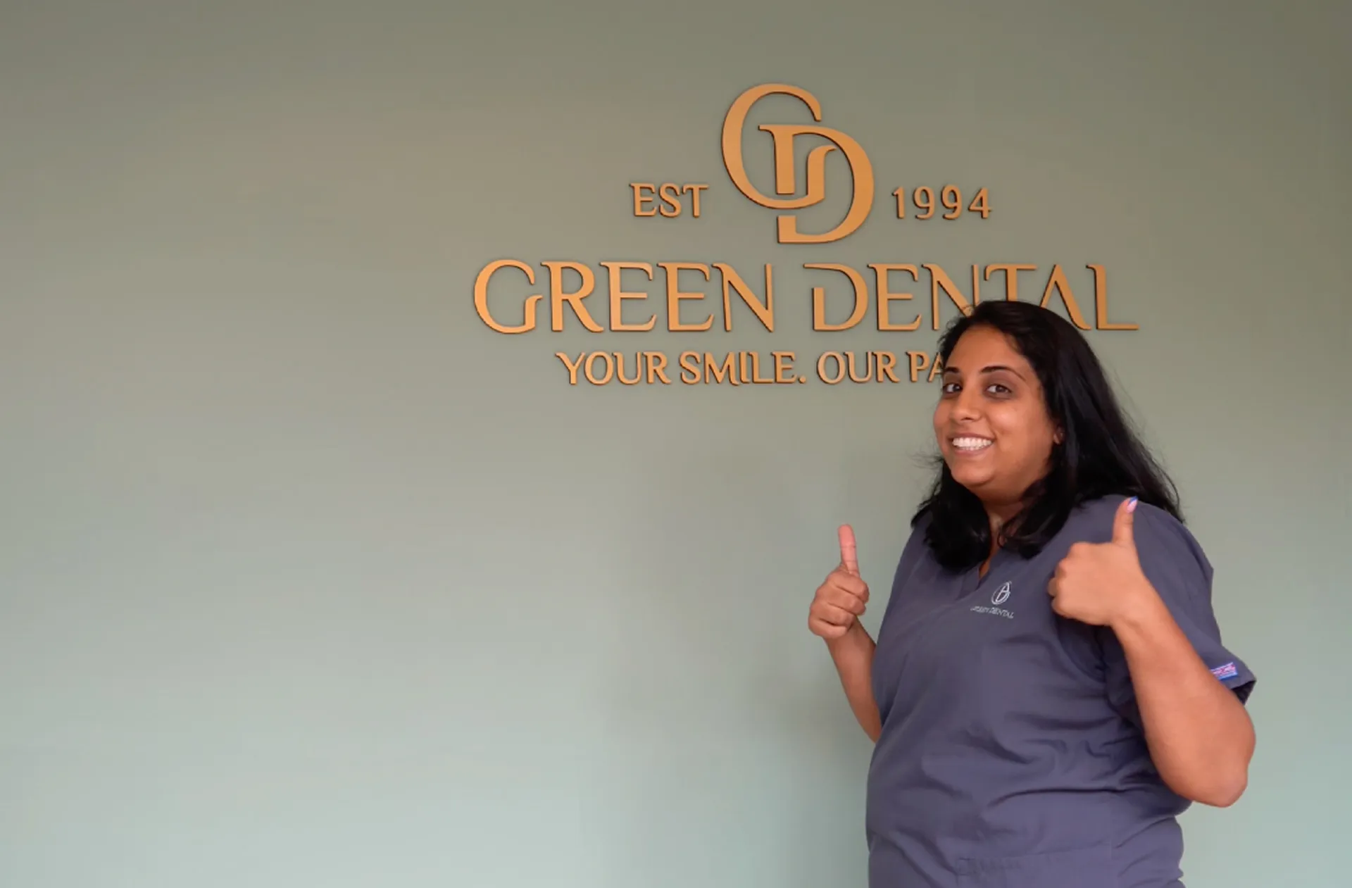 Smiling woman in dental scrubs showing thumbs up in front of a Green Dental sign on a green wall.