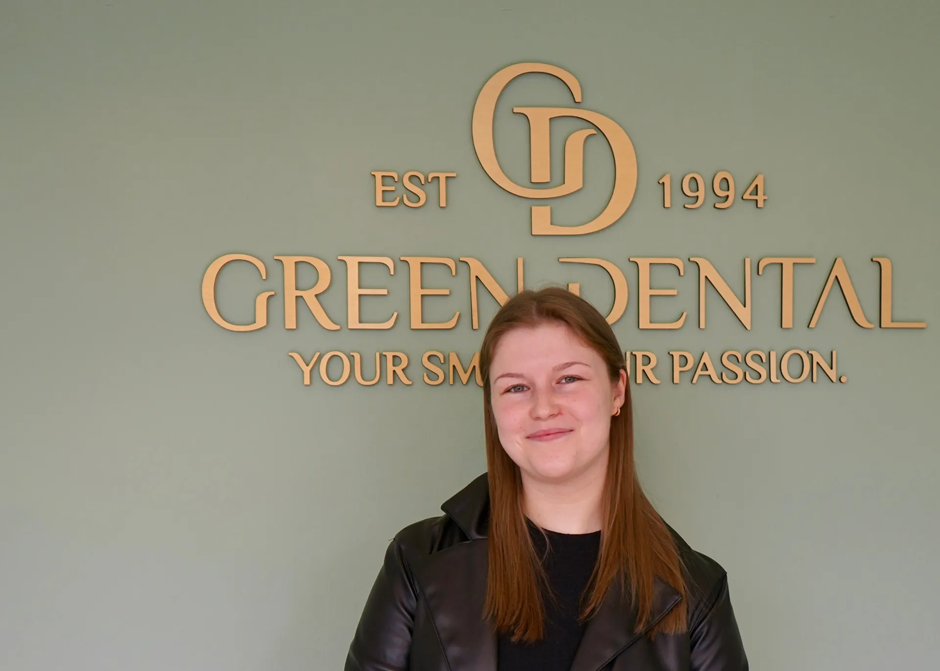 Smiling woman with straight brown hair wearing a black jacket standing in front of a green wall with Gold Green Dental logo and slogan.