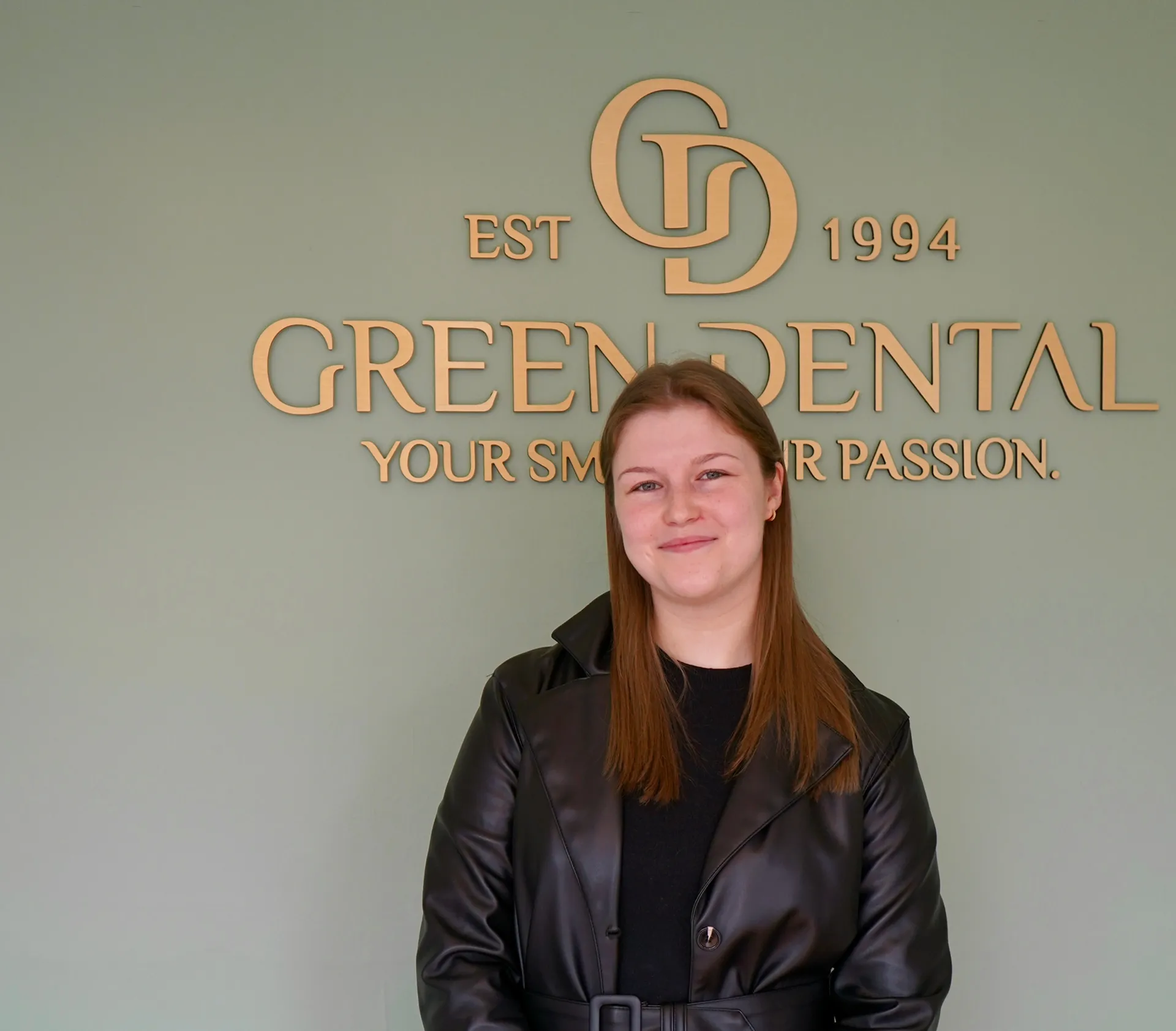 Smiling woman with long brown hair wearing a black leather jacket stands in front of a wall sign reading 'Green Dental, Your Smile, Our Passion, EST 1994.'