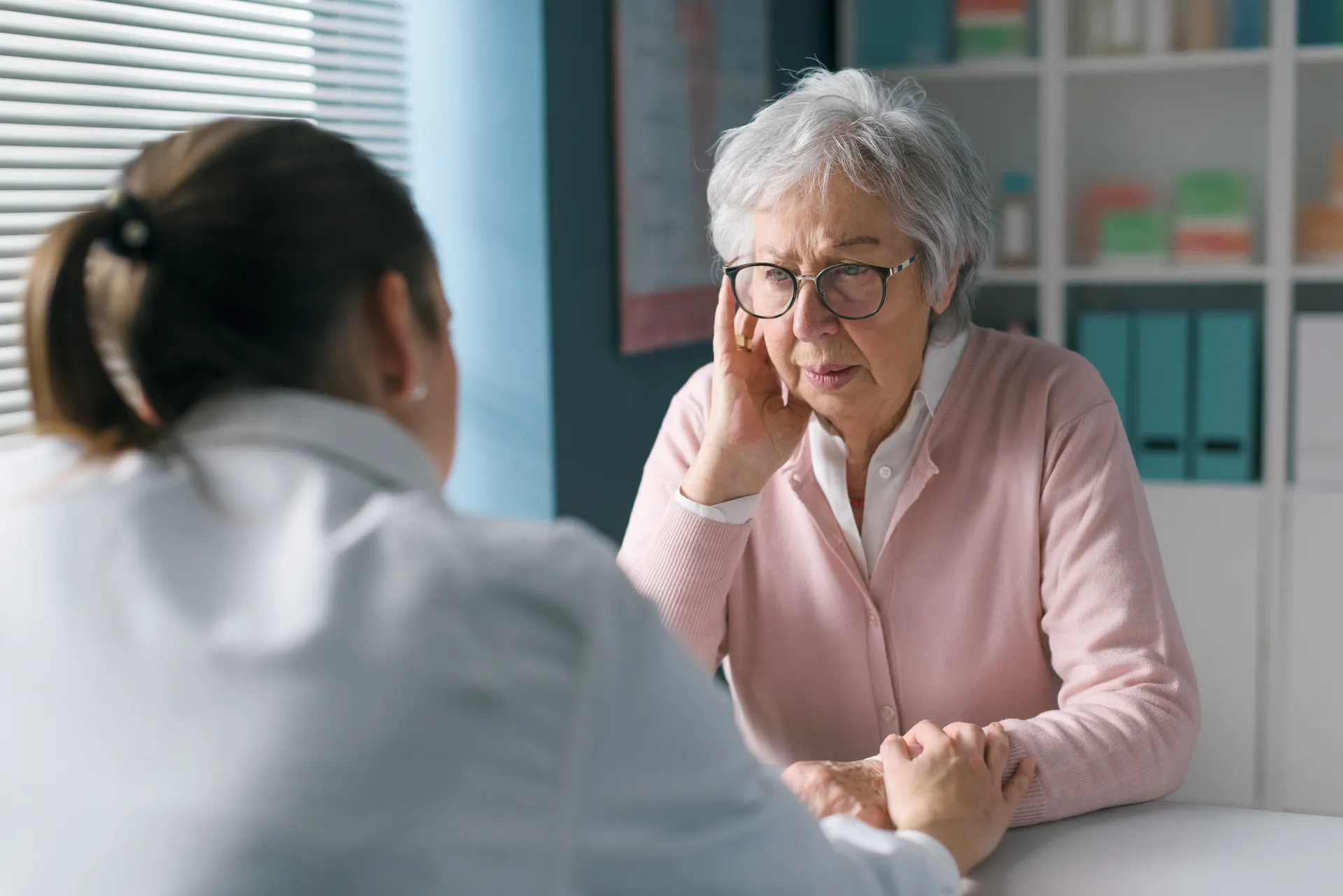Elderly woman in glasses holding her ear and looking concerned while talking to a doctor in an office.