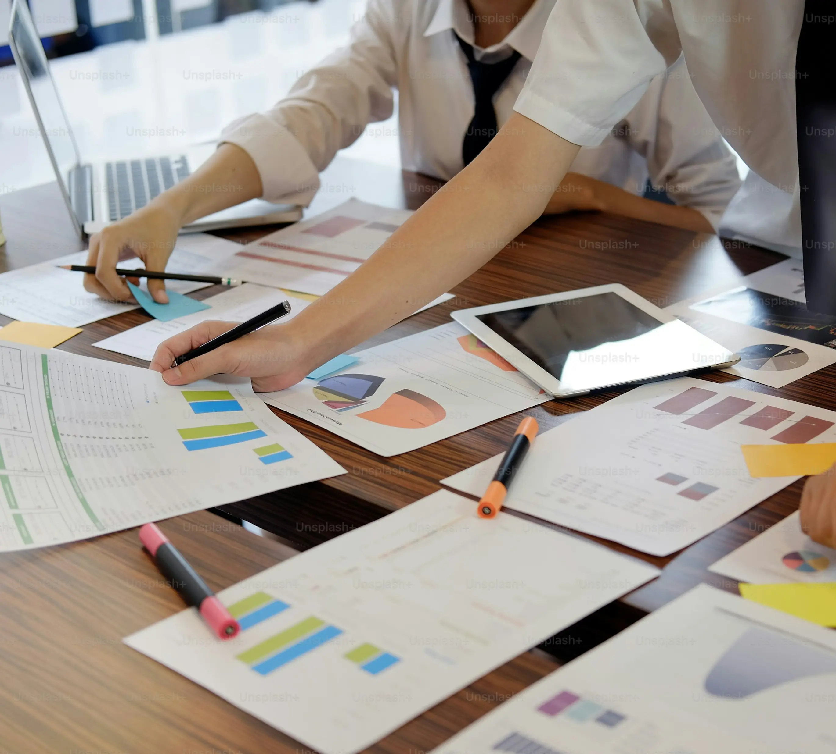 Two people in business attire reviewing printed charts and graphs on a wooden table with a tablet and laptop.