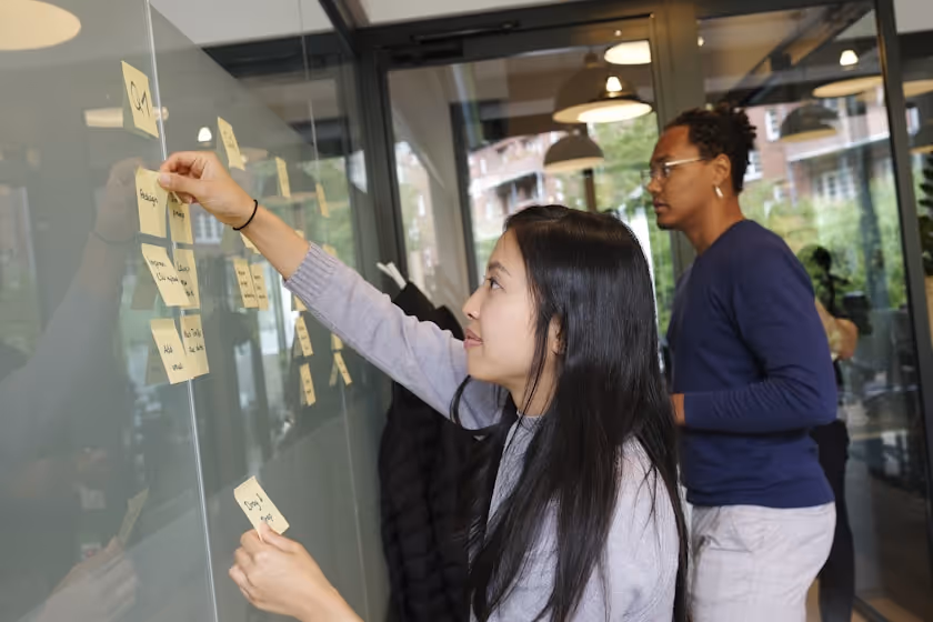 Two people attaching sticky notes on a glass wall in an office setting.