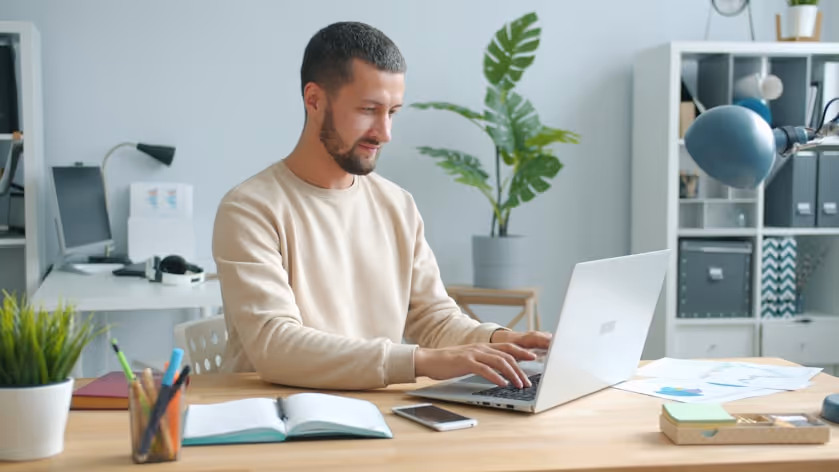 Man with short dark hair typing on a laptop at a wooden desk in a modern office space.