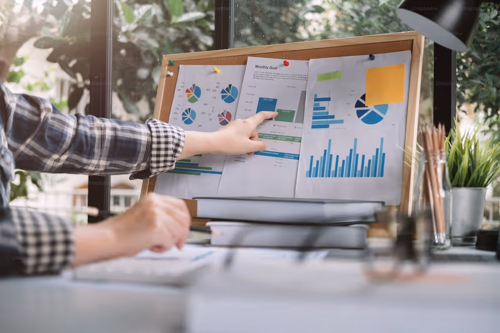 Person pointing at charts and graphs pinned on a corkboard in a sunlit office setting.
