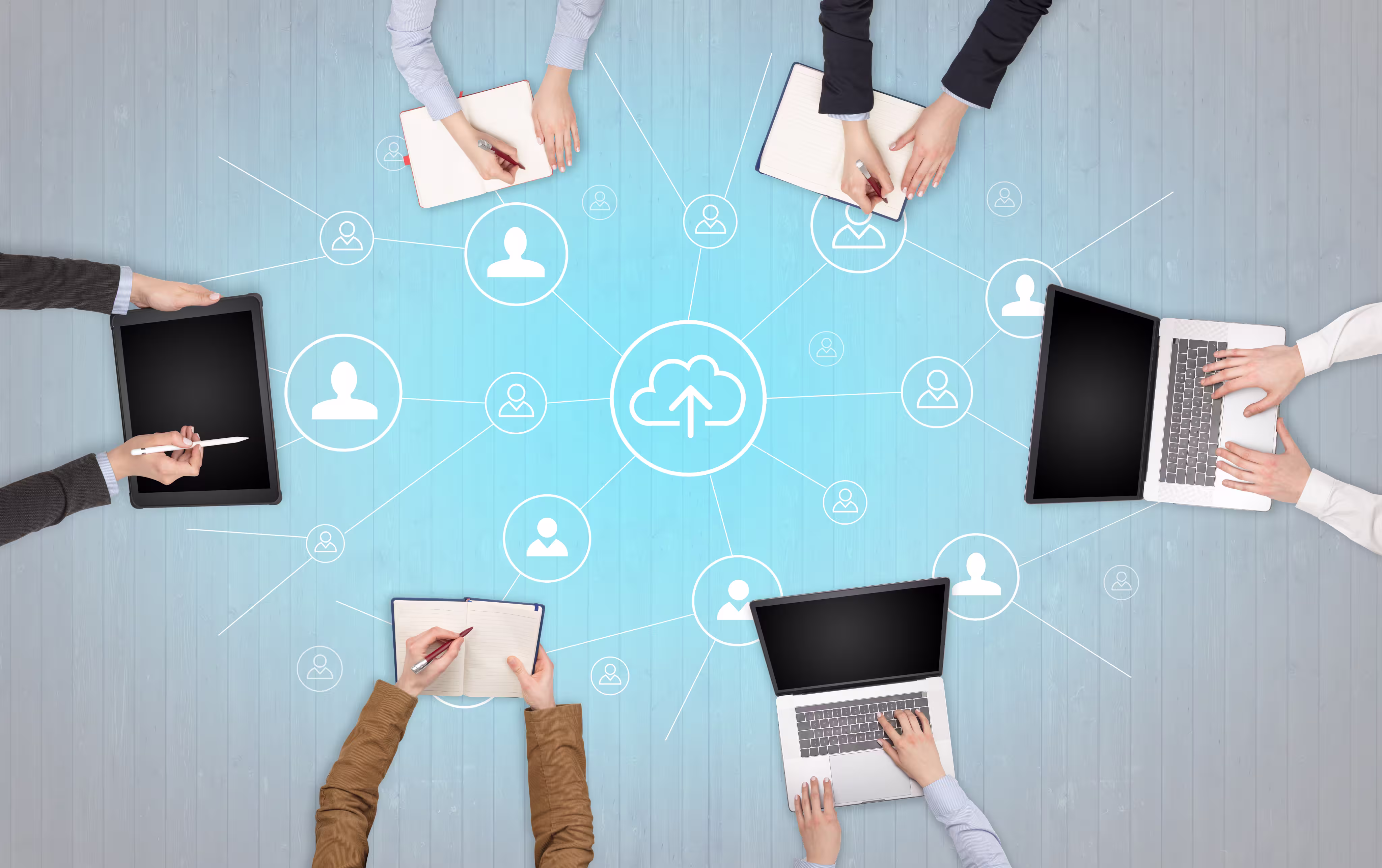 Overhead view of six people using laptops, tablets, and notebooks around a network diagram with cloud upload and user icons on a blue wooden table.