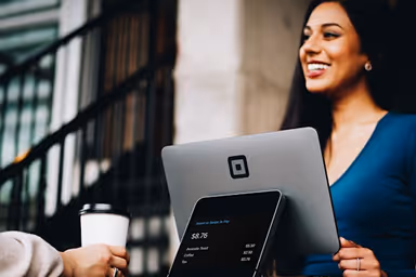 Smiling woman standing behind a cash register with a coffee cup nearby in a modern setting.