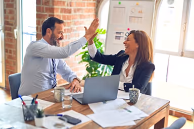Man and woman in business attire sharing a high-five across a table with a laptop and documents.