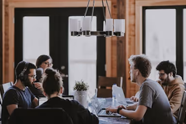 Group of five people working on laptops around a table in a cozy wooden room with large windows.
