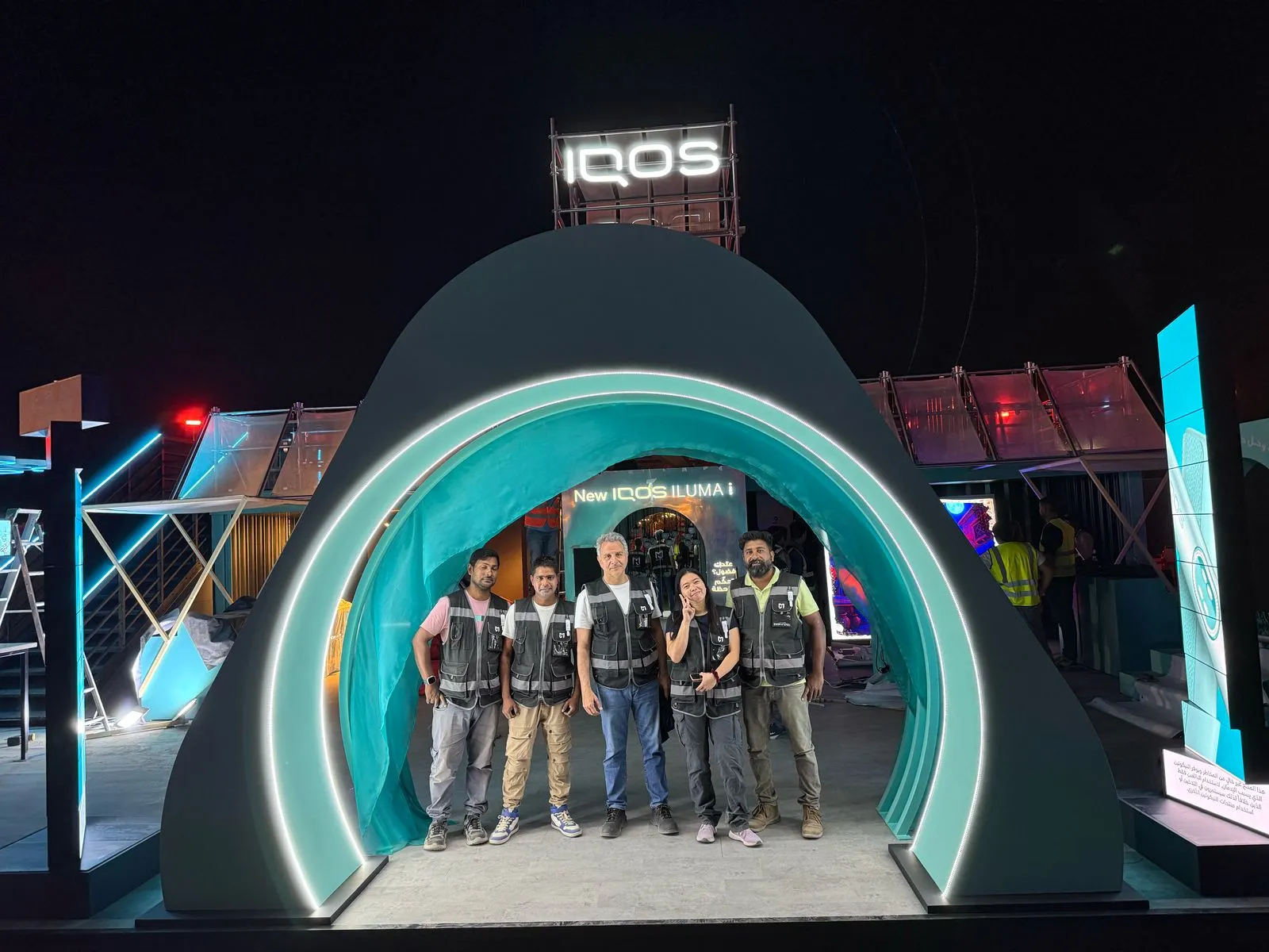 Five people wearing black vests stand smiling under a glowing archway with IQOS branding at night.