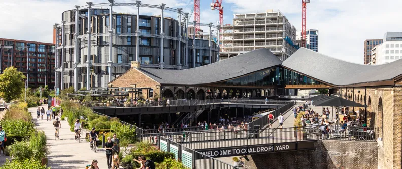 Outdoor view of Coal Drops Yard shopping and dining area with historic brick buildings, modern glass structures, and people walking and sitting at tables.