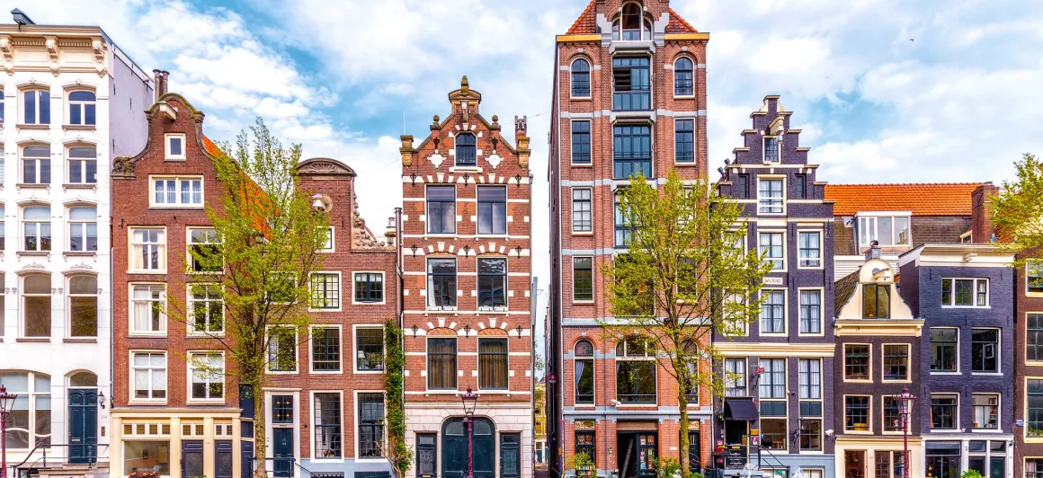 Row of traditional narrow Dutch canal houses with gabled roofs and trees in front under a partly cloudy sky.