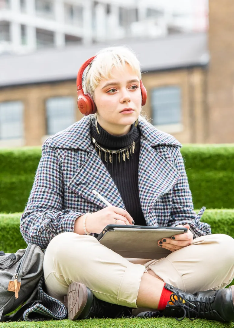 Young woman with short blonde hair wearing red headphones and a checkered coat, sitting cross-legged on grass while writing on a tablet with a stylus.