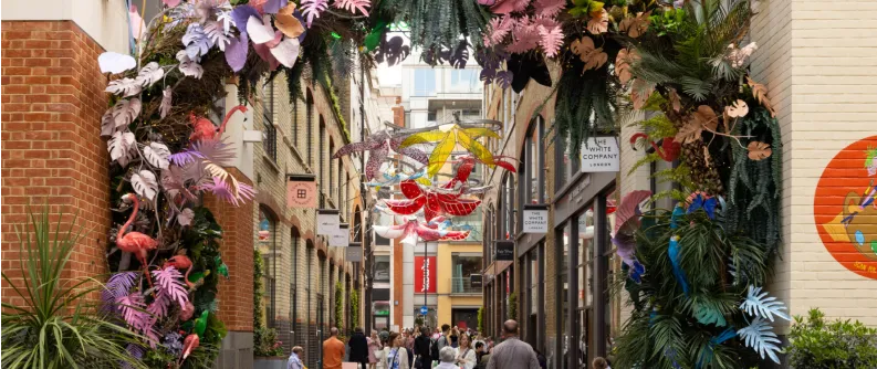 Busy urban alleyway decorated with colorful hanging sculptures and an arch of tropical plants and flowers.