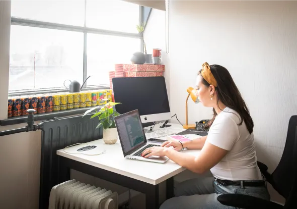 Woman with a headband working on a laptop at a bright desk near a window with a plant and computer monitor.