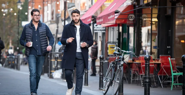 Two men walking on a city street holding coffee cups beside a parked bicycle and outdoor café seating.