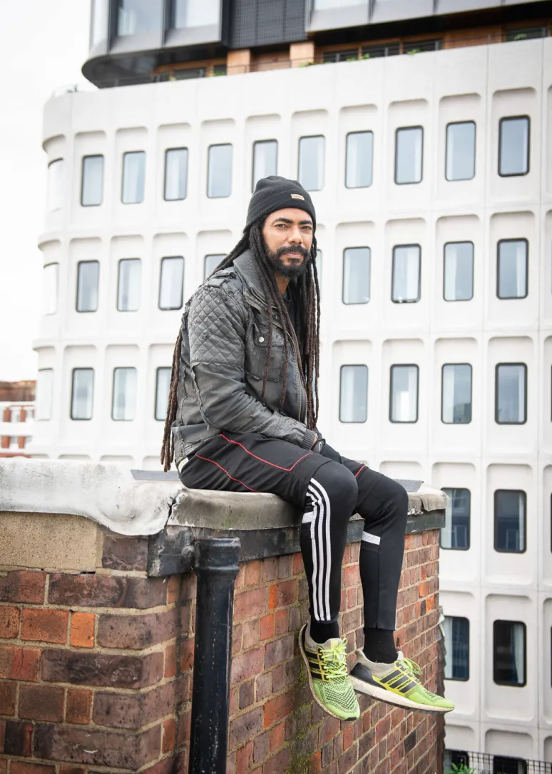 Man with long dreadlocks wearing a black beanie, quilted jacket, and athletic pants sitting on a brick ledge with a modern building in the background.