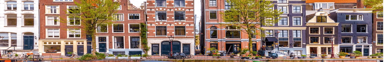 Row of traditional narrow brick canal houses with large windows and boats docked along the water in front.