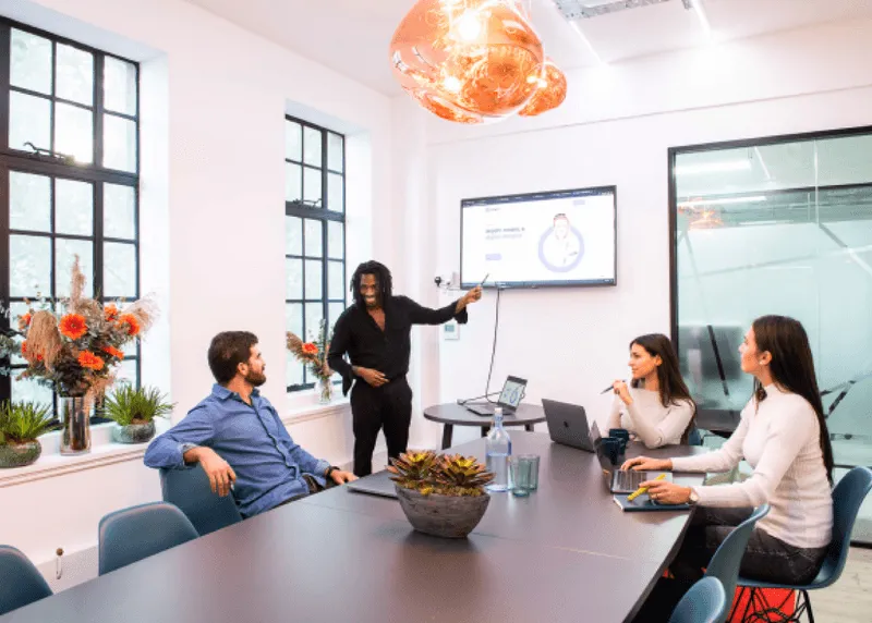 A man stands and points to a screen during a meeting with three seated colleagues in a modern conference room.