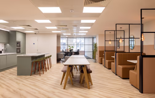 Modern office space with wooden floor, central tables with stools, and booth seating on the right separated by black metal frames.