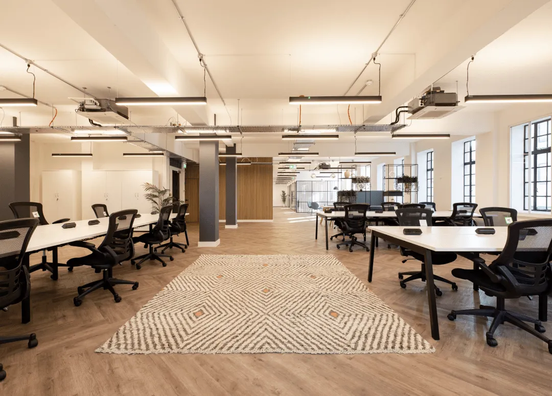 Spacious modern office interior with white desks, black ergonomic chairs, patterned rug, and large windows.