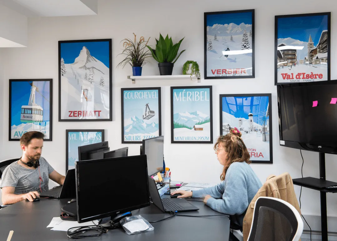 Two people working at desks with multiple computer monitors in an office decorated with framed vintage ski resort posters on the wall.