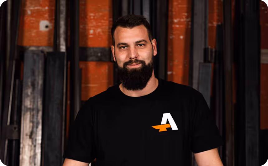 Mann mit Vollbart trägt ein schwarzes T-Shirt mit einem weißen und orangefarbenen Logo vor einer orange-schwarzen Wand.