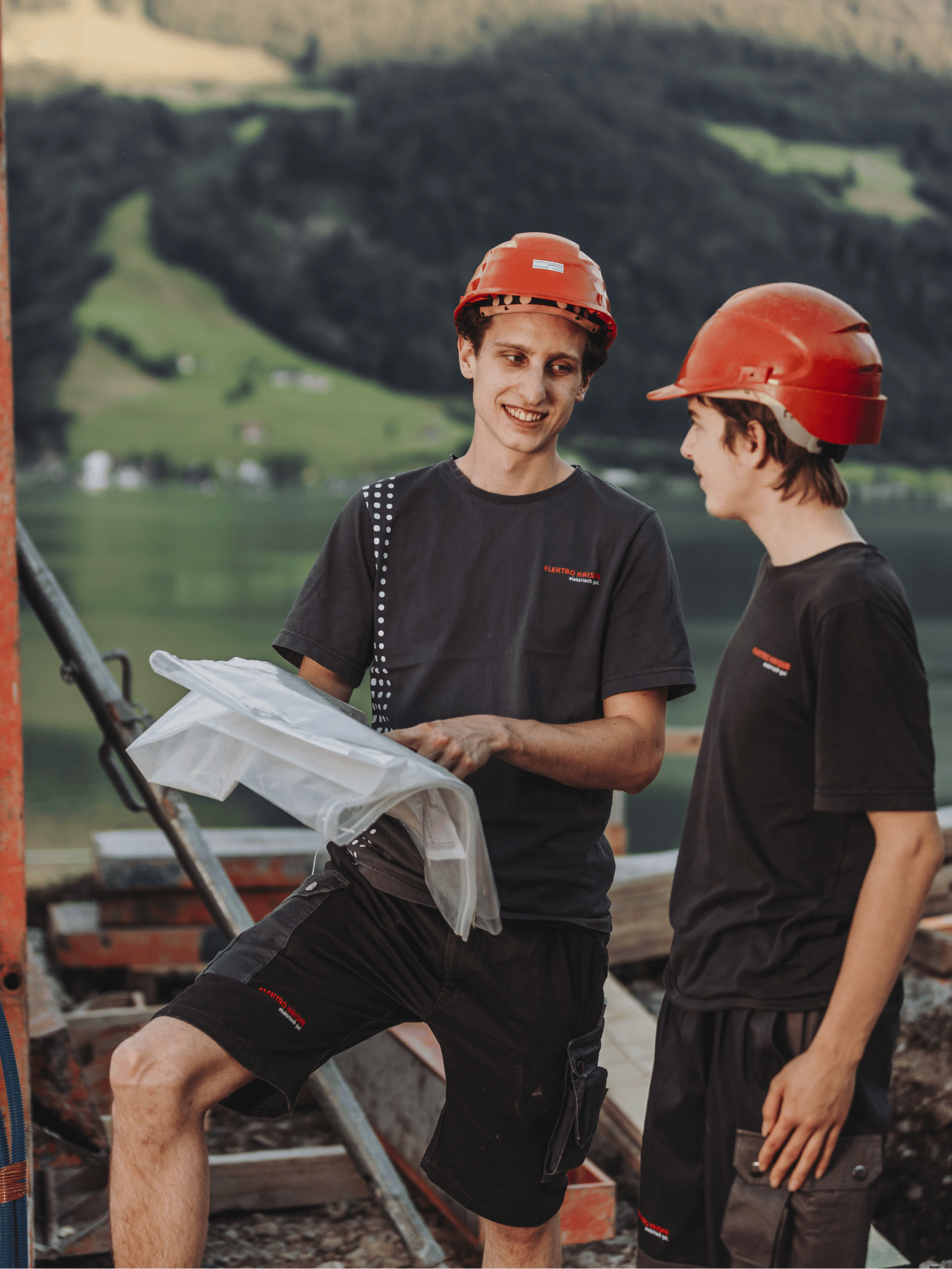 Zwei junge Bauarbeiter mit roten Schutzhelmen und dunklen Arbeitskleidern besprechen einen Bauplan vor einer Landschaft mit See und Bergen.