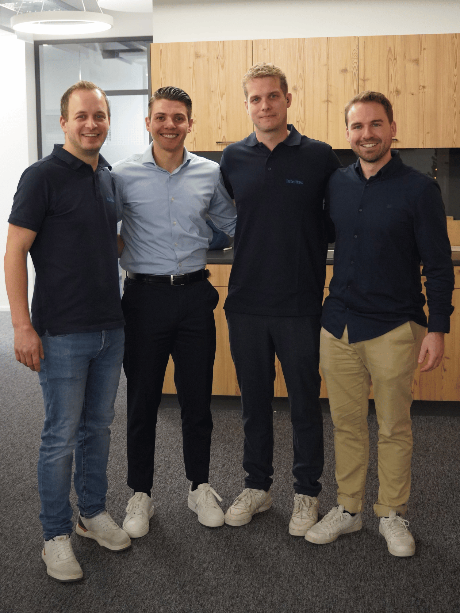 Vier Männer lächeln stehend vor einer Holzwand in einem modernen Büro.